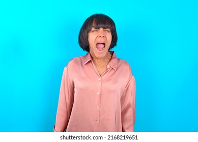 Stressful Young Brunette Woman Wearing Pink Silk Shirt Over Blue Background Screams In Panic, Closes Eyes In Terror, Keeps Hands On Head, Finds Out Terrified News, Can't Believe It.