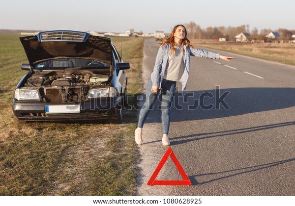Stressful\
young woman driver hitchhikes and stops cars, asks for help as have\
problem with brocken car, uses red triangle sign to warn drivers\
about stop. Auto pulled to side of country\
road