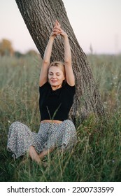 Stretching Under A Tree Blond Woman Sitting On Grass. She Is Wearing Skirt Pants And Black Shirt. Her Eyes Closed. Low Angle.