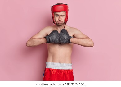 Strict angry unshaven male boxer holds hands together in boxing gloves ready for struggle wears protective helmet and shorts has bare torso isolated over pink background. Always ready for attack - Powered by Shutterstock