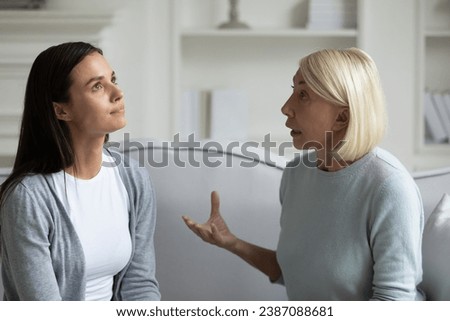 Strict anxious mature mother lecturing scolding annoyed grown daughter, sitting on sofa at home, parent and child quarrel, generations conflict, stubborn young woman ignoring angry senior mum