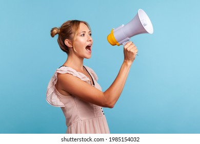 Strict blonde young adult woman with hair buns in summer dress loudly screaming at megaphone, making announce, protesting, wants to be heard. Indoor studio shot isolated on blue background.