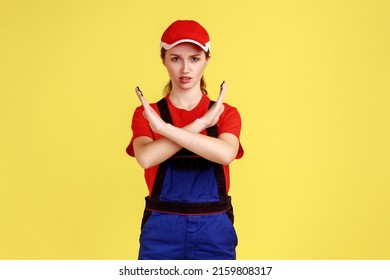 Strict Bossy Worker Man Standing And Looking At Camera With Serious Expression, Showing X Sign Gesture, Wearing Overalls And Red Cap. Indoor Studio Shot Isolated On Yellow Background.