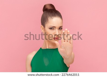 Strict bossy young adult woman standing with raised hand, showing stop gesture with palm, trying to stop aggressive actions, wearing green dress. Indoor studio shot isolated on pink background.