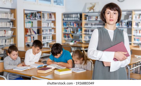 Strict Female Librarian Standing With Book In Hands In School Library On Background With Reading Children..