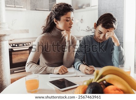 Strict mother checking how son doing homework at dining table at home