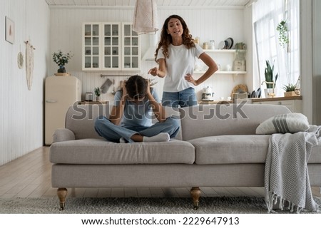 Strict mother scolding unhappy teenage daughter at home, mom lecturing teen girl, child sitting on sofa feeling afraid covering ears with hands. Parental harsh discipline, adolescent problem behavior 