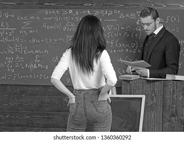 Strict Professor In Glasses Reading A Book At Rostrum. Female Brunette Student Holding Hands In Rear Jeans Pockets While Standing In Front Of Her Teacher.