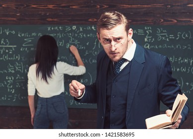 Strict Professor Looking At Students Over The Glasses While Holding Book And Piece Of Chalk In His Hands. Female Student Writing On Green Board.