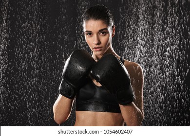 Strict Woman Doing Sports And Boxing In Gloves While Standing In Defense Position Under Rain Drops Isolated Over Black Background