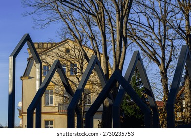 Striking black metal angular frames create a modern geometric installation in the foreground, framing a classic yellow historic building with tall bare trees and warm autumn light in a peaceful Polish town setting.