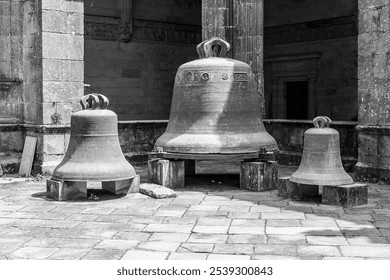 A striking black and white photograph showcasing three large, ornate bronze bells resting on wooden supports in a historic courtyard. - Powered by Shutterstock