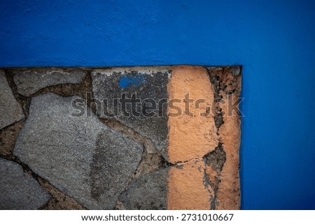 A striking close-up photograph capturing the sharp contrast between a smooth, bright blue painted wall and the rough, irregular texture of a rustic stone wall corner.