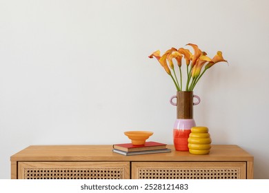 A striking floral centerpiece of orange calla lilies in a pink and orange ceramic vase, styled with a yellow decorative object and books on a wooden cabinet. - Powered by Shutterstock