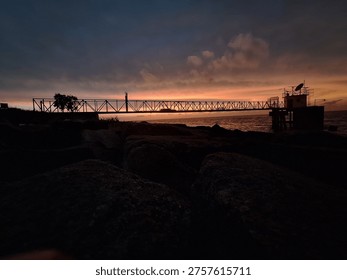 A striking, horizontal shot captures the dramatic silhouette of a long, metal industrial jetty extending over the calm ocean water during sunset or sunrise. The sky above is a vibrant display of warm orange and yellow hues near the horizon, contrasting sharply with the darker blue and grey clouds. The foreground is dark and rocky, providing a strong sense of depth and focus on the structure. This image evokes feelings of tranquility, industry, and the natural beauty of the coast at twilight. It is ideal for use in travel brochures, environmental campaigns, website backgrounds, or any project requiring a powerful, atmospheric coastal scene.