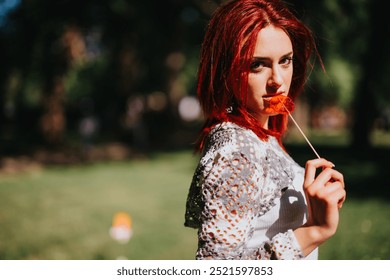 Striking image of a beautiful young redhead woman enjoying a lollipop outdoors in a lush green park on a bright sunny day. - Powered by Shutterstock