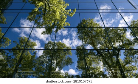 A striking image capturing the reflection of vibrant green trees and a cloudy blue sky on the glass exterior of a contemporary building, symbolizing nature and urban integration.