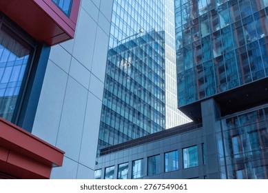 A striking low-angle photograph capturing the dynamic intersection of four architecturally distinct modern buildings under clear, natural daylight in a bustling urban environment.
This detailed, close-up shot offers a unique, abstract perspective on contemporary city architecture, showcasing a diverse array of materials and textures where four skyscrapers converge. From this viewpoint, the intricate composition highlights the geometric interplay of various building facades. In the left foreground, a modern structure with prominent red-orange metal accents frames a smooth, cool gray precast concrete facade with clean panel lines and deep blue-tinted windows. To the right, a multi-faceted glass skyscraper with textured, fractured-effect blue panes that reflect the sky and other elements creates a sharp corner. A second, central glass-and-steel high-rise, reflecting its surroundings with distinct square mullions, rises behind the others. Below this cluster, a darker building section with sequential dark metal louvered grilles and rows of large rectangular windows adds weight to the base. The lighting is crisp natural daylight, enhancing the cool blue reflections in the glass and the warm tone of the red accents, while showing a glimpse of a nearby crane at the top. The image is a complex, geometric tapestry of urban development, offering an eye-catching pattern of vertical lines, grids, and material contrasts.