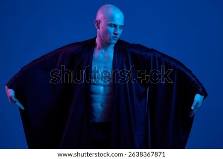 Striking man in dramatic pose, showcasing a toned physique, dressed in flowing black robe, set against a captivating blue background.