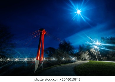 A striking night scene featuring a suspension bridge illuminated by the moonlight. The bridge’s red support contrasts against the deep blue sky, with reflections glimmering on the calm river below. - Powered by Shutterstock