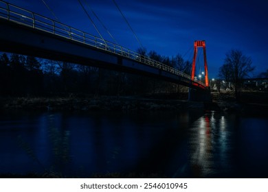 A striking night scene featuring a suspension bridge illuminated by the moonlight. The bridge’s red support contrasts against the deep blue sky, with reflections glimmering on the calm river below. - Powered by Shutterstock