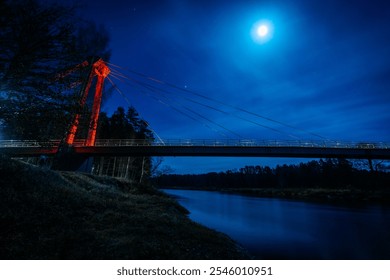 A striking night scene featuring a suspension bridge illuminated by the moonlight. The bridge’s red support contrasts against the deep blue sky, with reflections glimmering on the calm river below. - Powered by Shutterstock