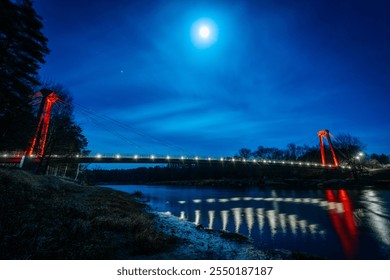 A striking night scene featuring a suspension bridge illuminated by the moonlight. The bridge’s red support contrasts against the deep blue sky, with reflections glimmering on the calm river below. - Powered by Shutterstock