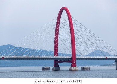 Striking red cable-stayed bridge spanning across blue water with mountains in background, modern architectural design with symmetrical cables and elegant arch structure.