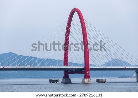 Striking red cable-stayed bridge spanning across blue water with mountains in background, modern architectural design with symmetrical cables and elegant arch structure.