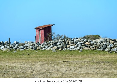 A striking red shed stands alone against a clear blue sky, bordered by a neatly stacked stone wall, showcasing the tranquility of a rural setting - Powered by Shutterstock