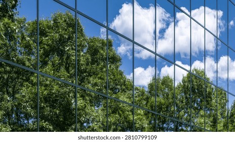 A striking reflection of vibrant green trees and fluffy white clouds in a bright blue sky is captured on the glass facade of a contemporary building.