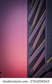 A striking, split-composition photograph viewed from a high-angle perspective. The left half of the frame is dominated by a deep, glowing pink and magenta sunset sky at dusk. The entire scene is bathed in this saturated, soft light. On the right, the geometric facade of a modern hotel building recedes into a dense, repeating grid. Multiple stories of identical glass-railed balconies form a powerful perspective line, each illuminated by warm, golden-yellow LED strip lights. The lower glass curtain wall is dark, reflecting deep red and pink tones. The clean lines, repeating forms, and the juxtaposition of organic color and man-made structure define the composition. The focus is sharp across the building details and the distant windows. Diffuse natural light of dusk creates deep saturation. There are no people visible, and no signs of specific cities or countries. The texture of the tree and the geometric lines of the building are prominent.