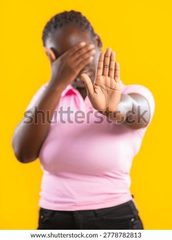 A striking studio image features a young Black woman against a vivid yellow background. She is captured in a dynamic pose of refusal, covering her eyes with one hand while extending the other palm toward the viewer in a firm 