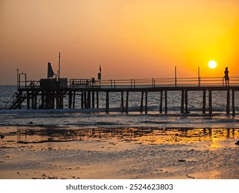 A striking sunset scene with a pier and figures walking on it. The sun casts a warm glow over the water, creating a peaceful atmosphere and enhancing the beauty of the coastal landscape. - Powered by Shutterstock