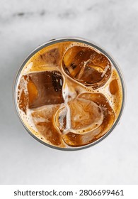 A striking top-down view looking directly into a glass of refreshing iced coffee or an iced latte. Large, clear ice cubes are suspended in the rich espresso, surrounded by creamy swirls of milk and a delicate golden-brown coffee foam. The circular glass is centered against a clean, light marble surface, creating a beautiful minimalist aesthetic. This crisp, bright commercial beverage photography is perfect for cafe menus, specialty coffee shop branding, and summer refreshment advertising.