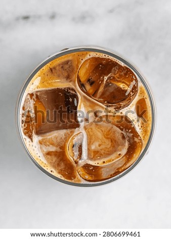 A striking top-down view looking directly into a glass of refreshing iced coffee or an iced latte. Large, clear ice cubes are suspended in the rich espresso, surrounded by creamy swirls of milk and a delicate golden-brown coffee foam. The circular glass is centered against a clean, light marble surface, creating a beautiful minimalist aesthetic. This crisp, bright commercial beverage photography is perfect for cafe menus, specialty coffee shop branding, and summer refreshment advertising.