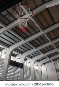 A striking vertical view capturing the intersection of mid-century architecture and sports culture. The red-and-white woven net takes center stage beneath the dramatic curve of massive white concrete ribs, framed by towering latticed windows that bathe the expansive interior in a gentle, atmospheric glow.