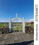 A striking white archway with a cross marks the entrance to a historic Icelandic cemetery, framed by a scenic coastal landscape and a deep blue sky, Iceland.