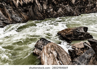 A striking wide-angle view highlighting the dramatic geological architecture of Tiger Leaping Gorge in Yunnan Province, China. The raging, olive-green Jinsha River tears through the canyon floor, churning into white rapids past massive foreground boulders. Above the torrent, the sheer canyon wall displays magnificent, steeply inclined metamorphic strata, offering a vivid cross-section of the immense tectonic forces that shaped this legendary southwestern Chinese landscape.
