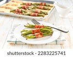 String beans wrapped in a roasted slices of bacon in a ceramic plate on a white wooden table, close up.
