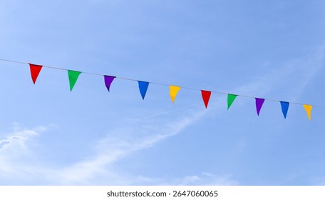 String of colorful triangular pennant or bunting flags hanging diagonally against a clear blue sky, symbolizing celebration, festivity, and outdoor events. Minimalist composition with vibrant tones.