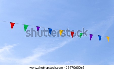 String of colorful triangular pennant or bunting flags hanging diagonally against a clear blue sky, symbolizing celebration, festivity, and outdoor events. Minimalist composition with vibrant tones.