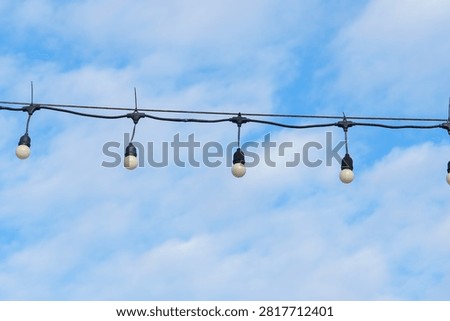 String light bulbs hanging on wire against bright blue sky with soft clouds, creating calm and peaceful outdoor atmosphere