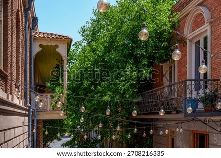 String lights crisscross between brick houses and ornate balconies above a narrow leafy side street.