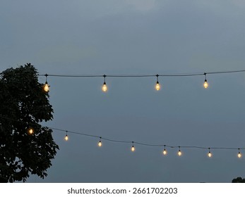 String lights glowing at dusk, hanging above an outdoor space with a tree silhouette, creating a cozy and inviting evening atmosphere.