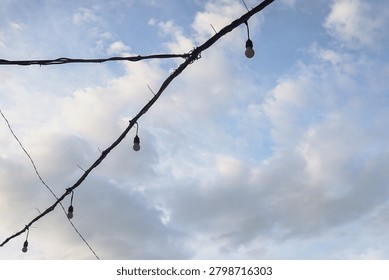 String lights hang against a soft, cloudy sky. Each bulb casts a gentle glow, inviting warmth. Faint wires stretch across the frame, adding texture