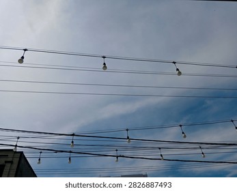 String lights with light bulbs hanging on wires against a bright blue sky with soft white clouds, low angle view of outdoor cafe or event decoration.
