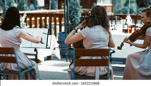 String Orchestra Performing Classical Music Outside Summer Terrace Three Wemen Wearin Lilliac Dresses And Holding Violins Shot From Behind.