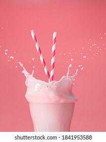 striped straws in a glass of splashing strawberry milkshake isolated on pastel pink color backdrop