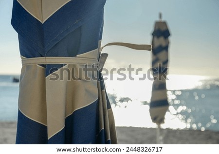 A striped umbrella is on the beach. The sky is blue and the water is calm. The umbrella is tied to a pole